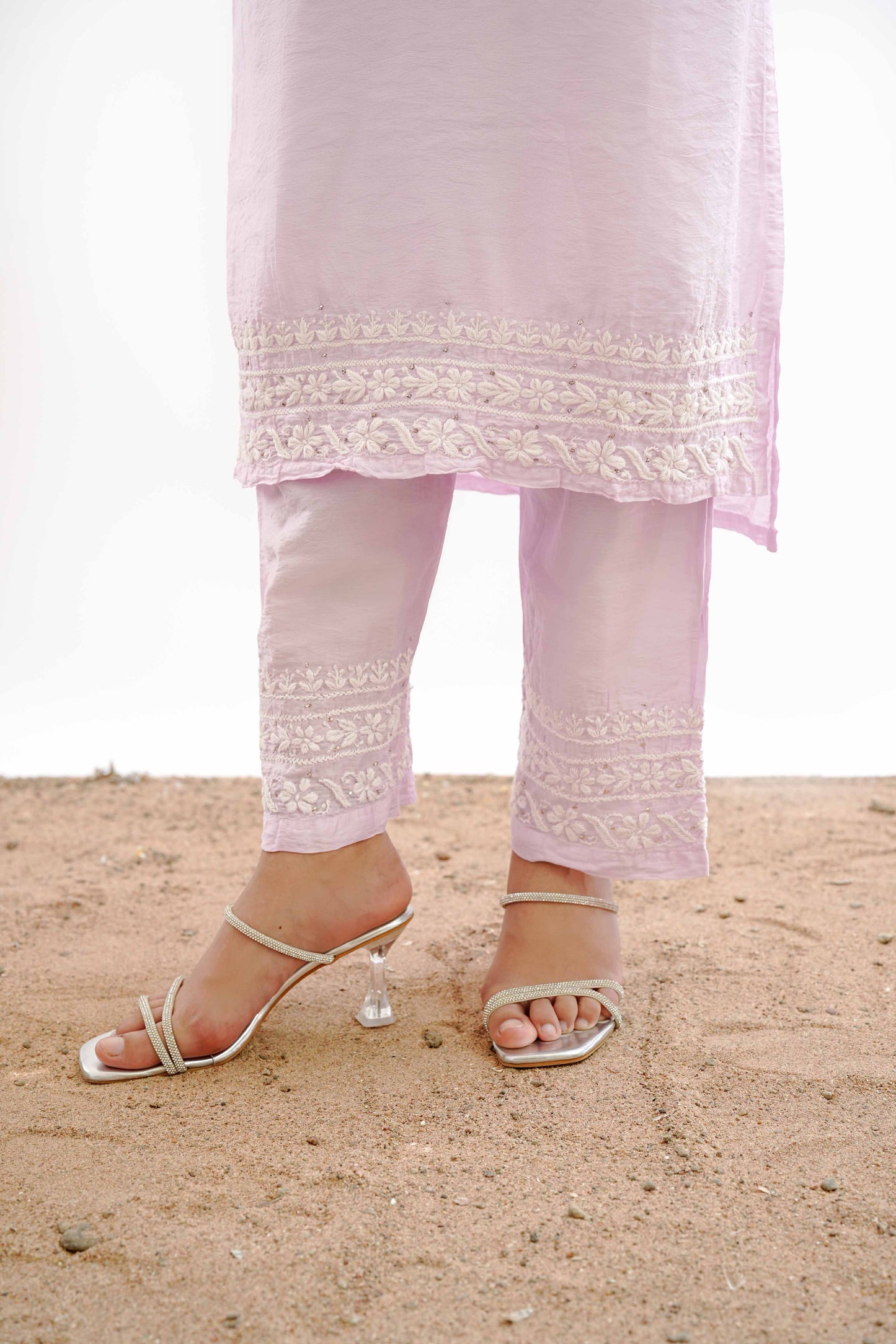 A person wearing a light pink traditional outfit with white embroidered borders on the pants and tunic, paired with silver heeled sandals, standing on sand against a white background.IshqaraPastelPinkMukaishChanderiKurtaSet3