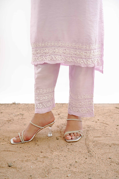 A person wearing a light pink traditional outfit with white embroidered borders on the pants and tunic, paired with silver heeled sandals, standing on sand against a white background.IshqaraPastelPinkMukaishChanderiKurtaSet3
